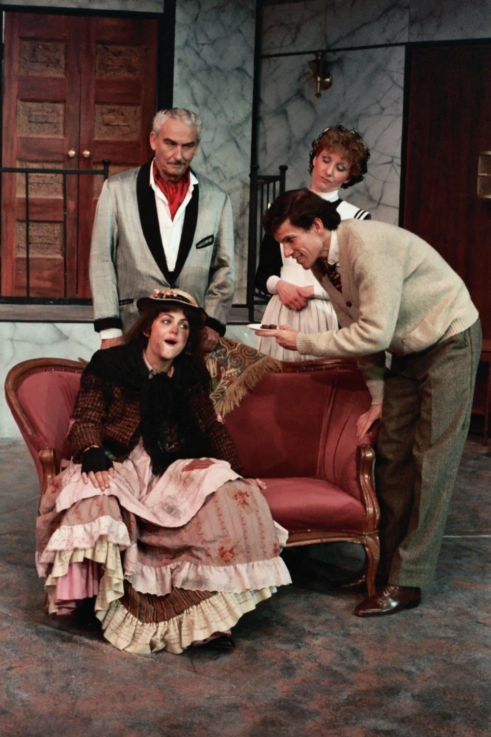 Theatrical scene with four actors in period costumes; a man offers chocolates to a woman sitting on a sofa; two people stand behind observing.
