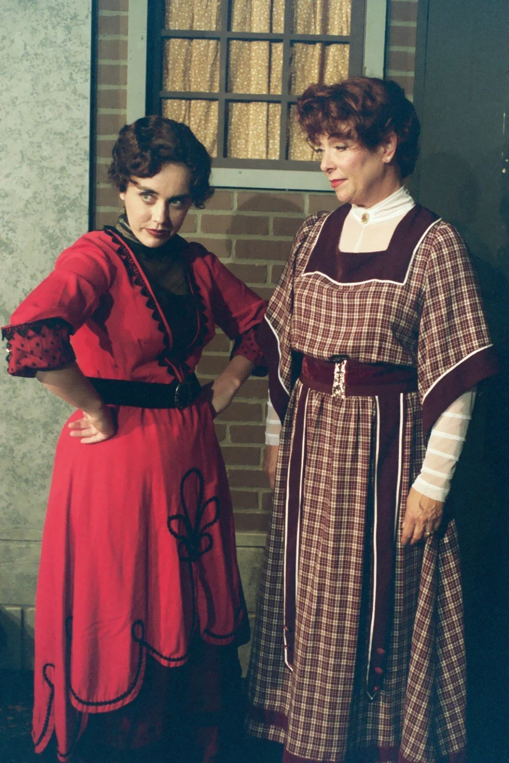 Two women in period costumes stand in front of a brick wall, with one wearing a red dress and the other in a plaid dress.