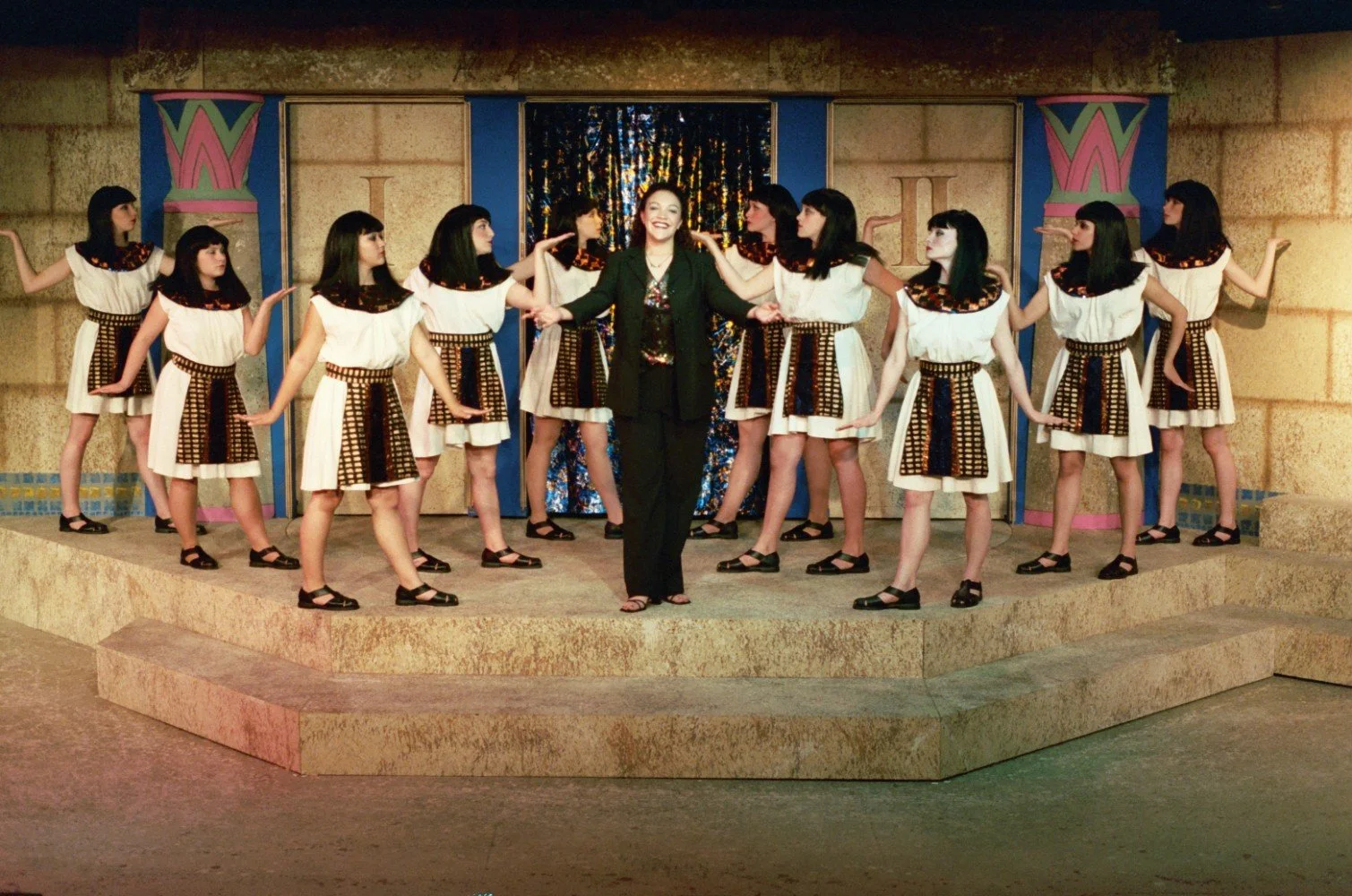 A group of performers on stage in Egyptian-inspired costumes, surrounding a person in modern clothing. The stage has Egyptian-style decor.
