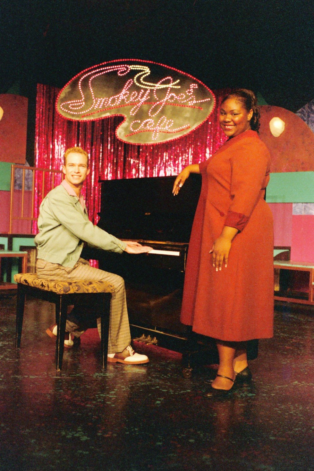 Two people on stage with a piano; one is sitting at the piano, and the other is standing, with a "Smokey Joe's Café" sign in the background.