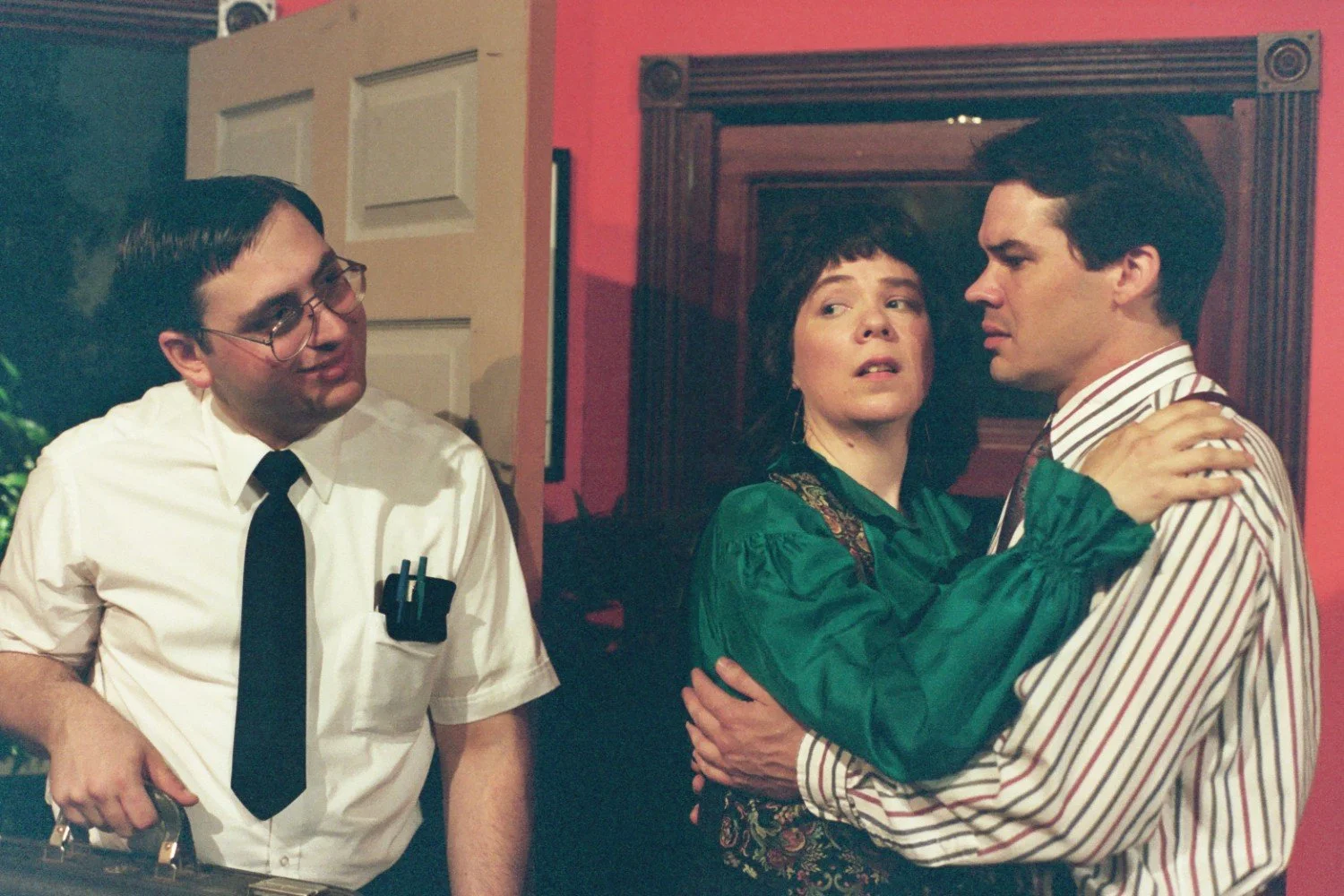 Three people indoors, one holding a briefcase and wearing a tie, the other two in an embrace, looking concerned.