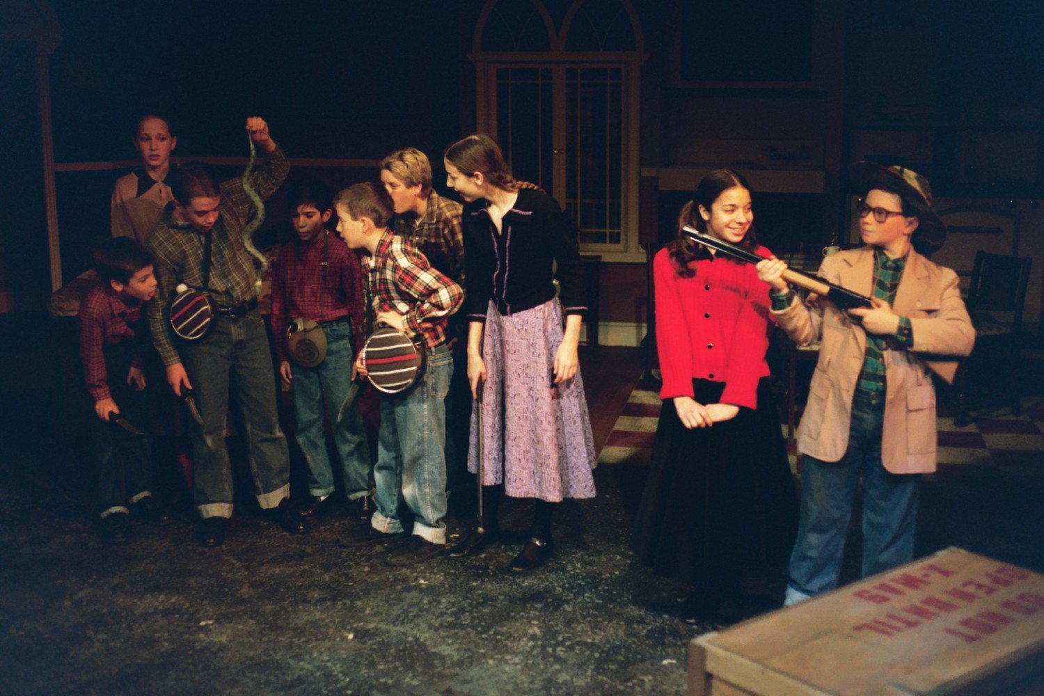 A group of children in costume on stage during a theatrical performance. One child holds a snake, others wear plaid shirts and overalls, and a girl in a red coat stands next to a boy with a hat and rifle. There's a wooden crate in the foreground.