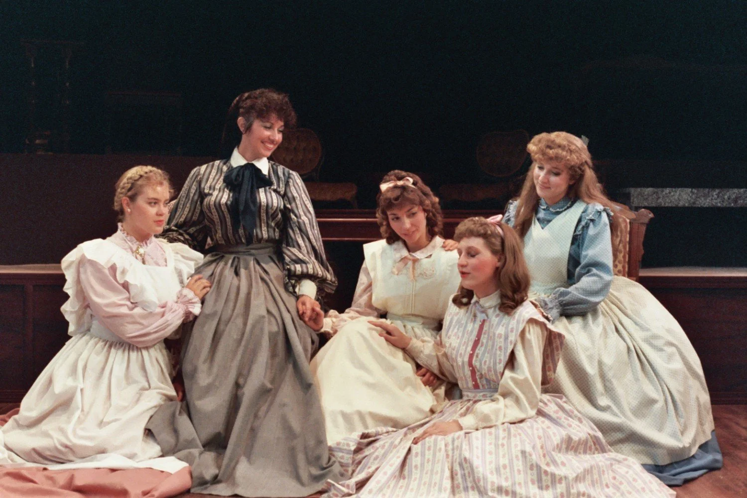 Five women in Victorian-era dresses seated together on a stage, appearing to perform a scene from a play.
