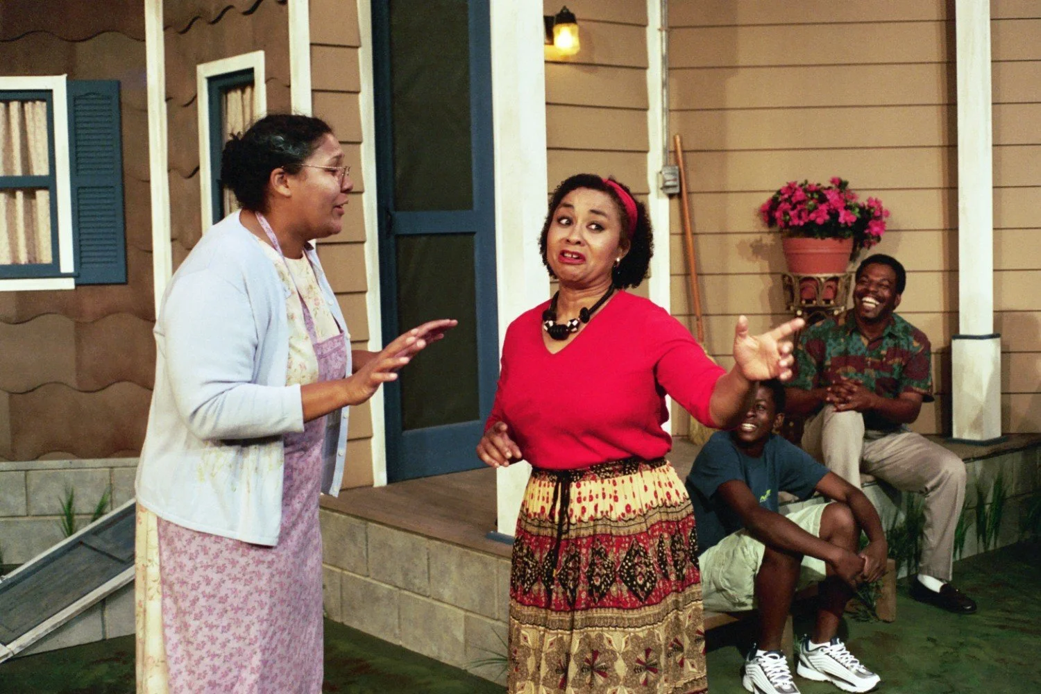 Two women in conversation in front of a house, with two men sitting nearby. One woman wears an apron, the other a red top and patterned skirt. A flower pot is visible.
