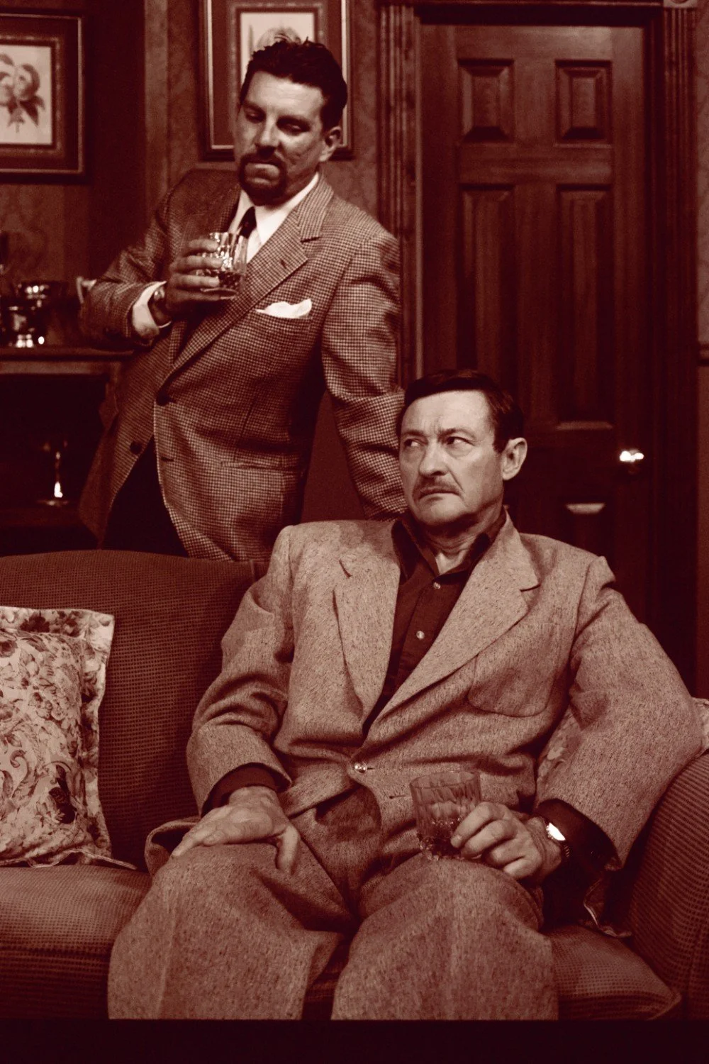 Two men in vintage suits holding drinks in a dim, wood-paneled room.