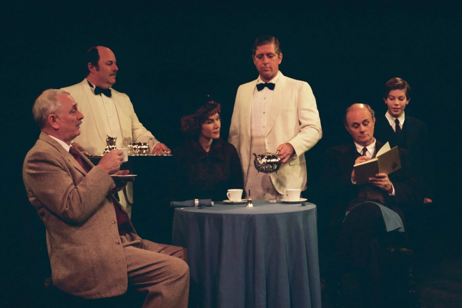 Scene of actors in vintage-style costumes portraying a tea party, with one person pouring tea, others seated and holding teacups, and one reading a book.