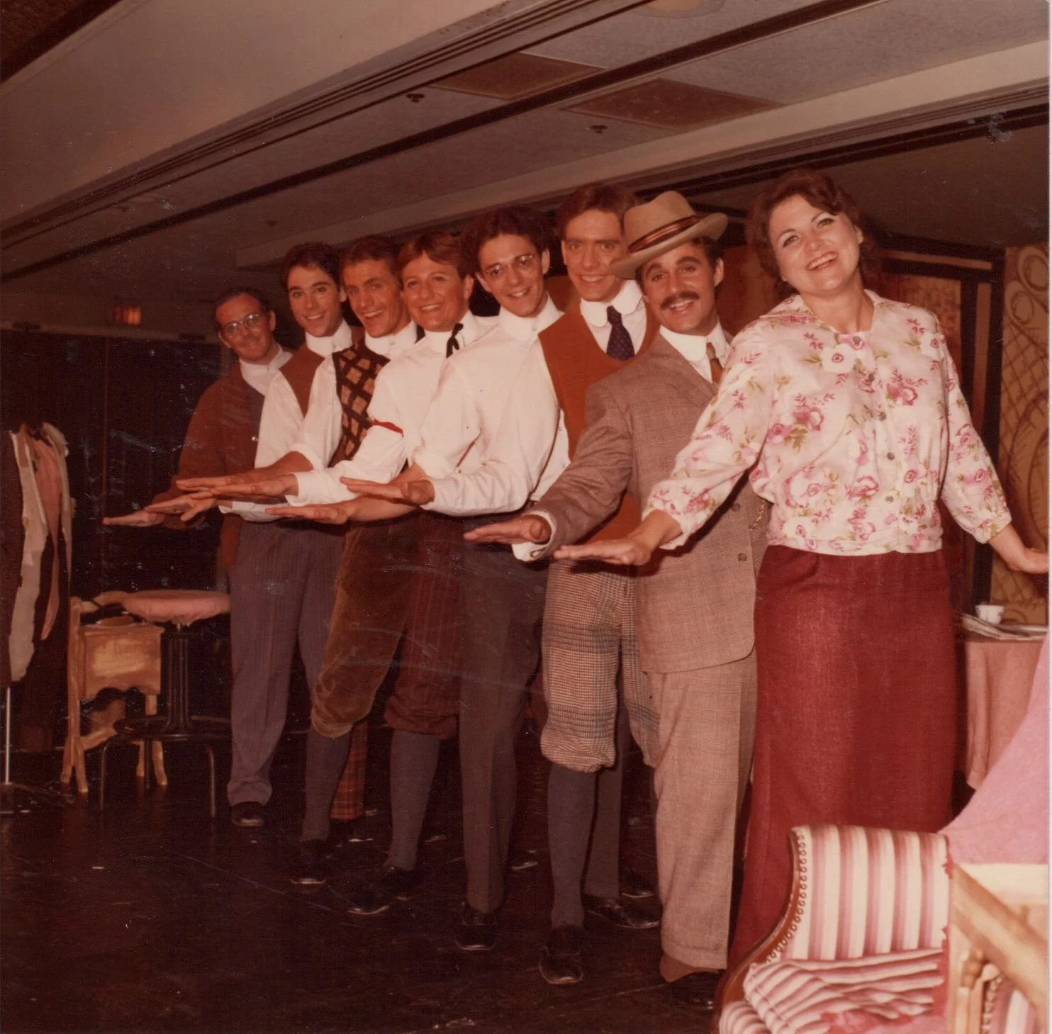 A group of eight people standing in a line, dressed in vintage-style clothing, posing with extended hands in a theatrical setting.