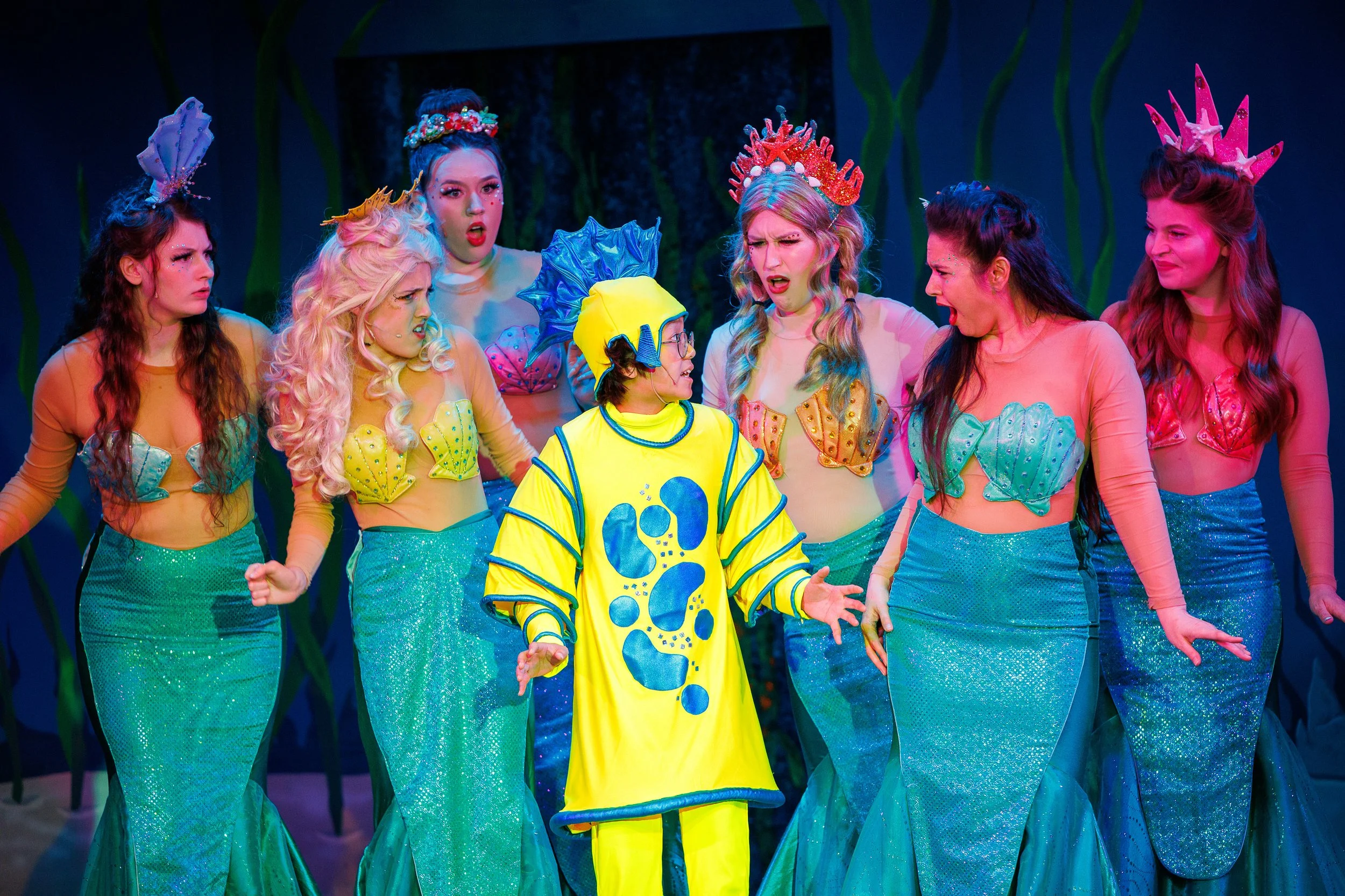 Theatrical performance with six actors dressed as mermaids and one as a fish, on stage.