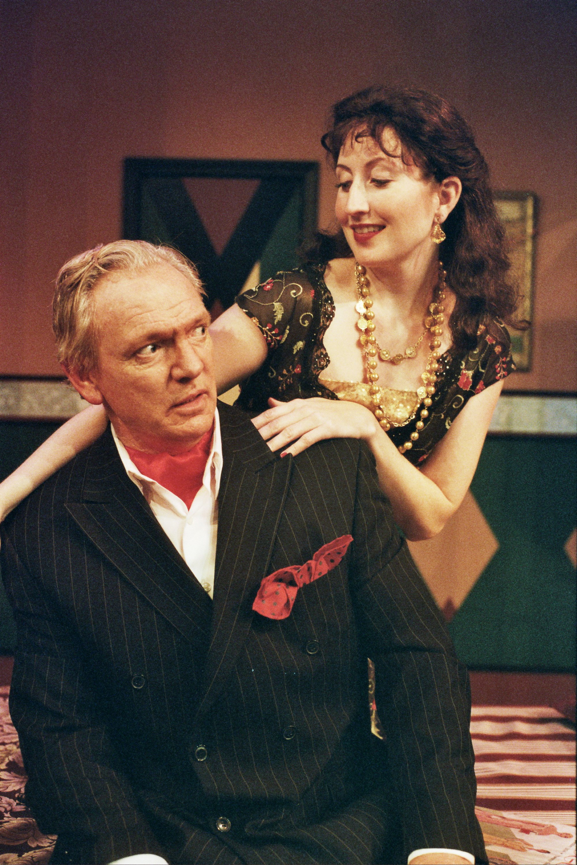 A woman in a black dress with colorful details and gold jewelry stands behind a man in a pinstripe suit. She is smiling and has her hands on his shoulders, while he looks surprised or concerned.