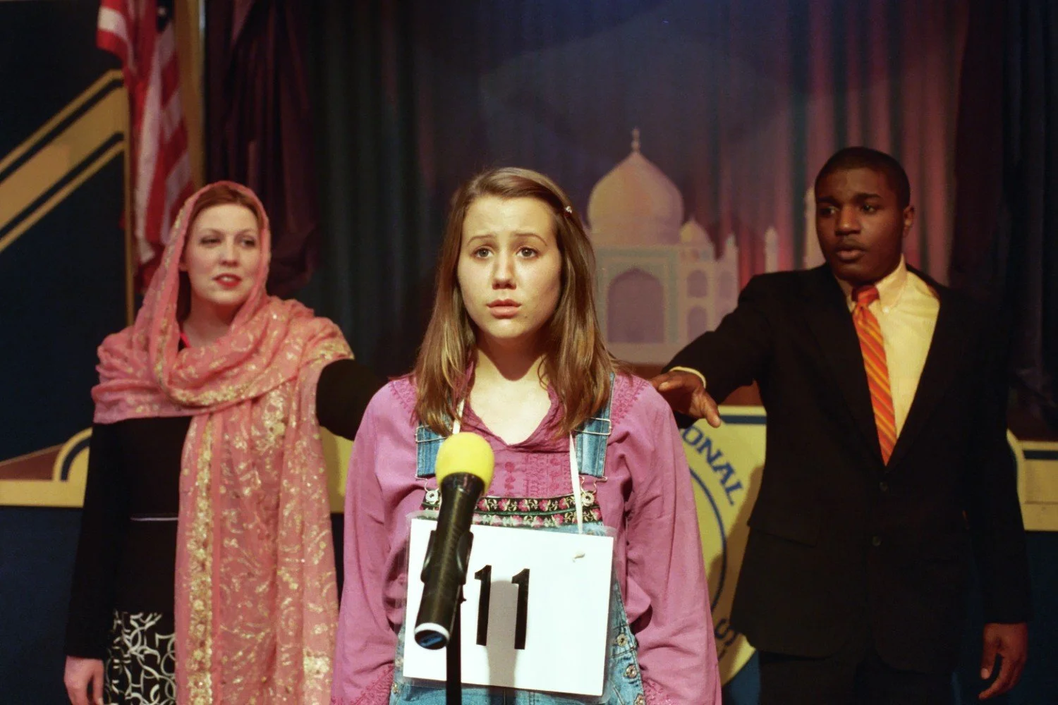 A girl stands in front of a microphone wearing a "11" number tag, flanked by two adults, one in a pink headscarf and the other in a suit, during a spelling bee event.