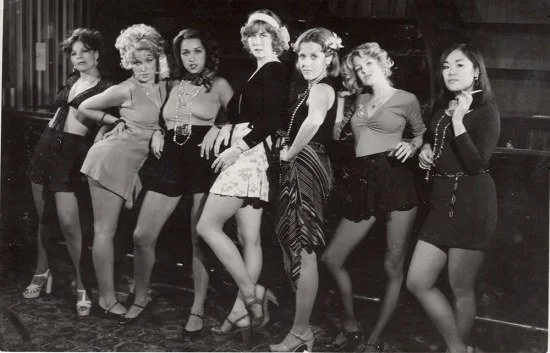 Black and white photo of seven women lined up, dressed in vintage 1960s fashion, wearing short dresses or skirts and heels.