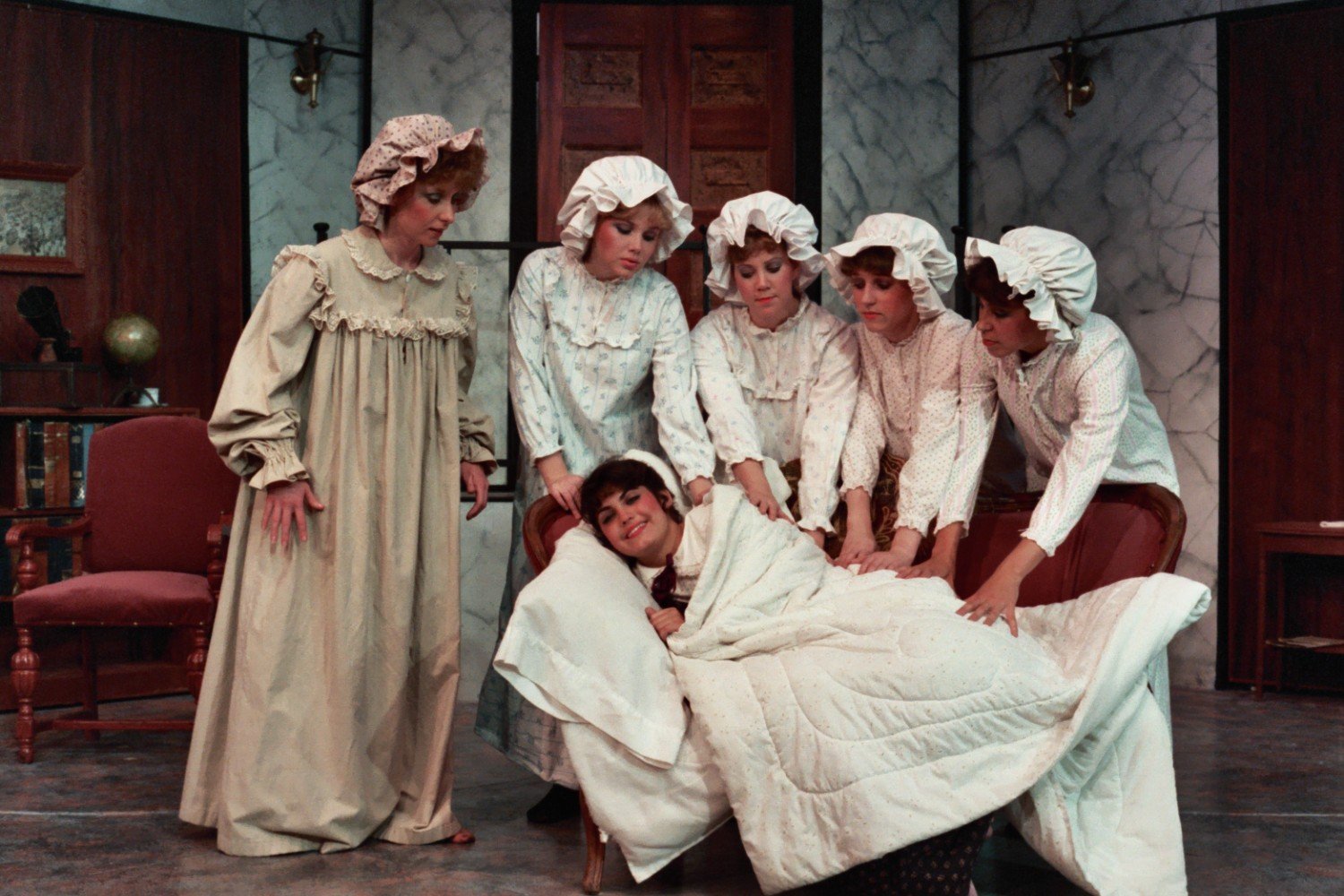 People in period costumes on stage, with six women wearing nightgowns and bonnets standing around a person lying on a couch, covered in a white blanket.