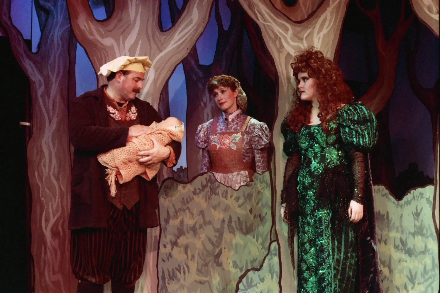 Theatrical performance with three actors in forest scene, one holding a baby wrapped in a blanket.