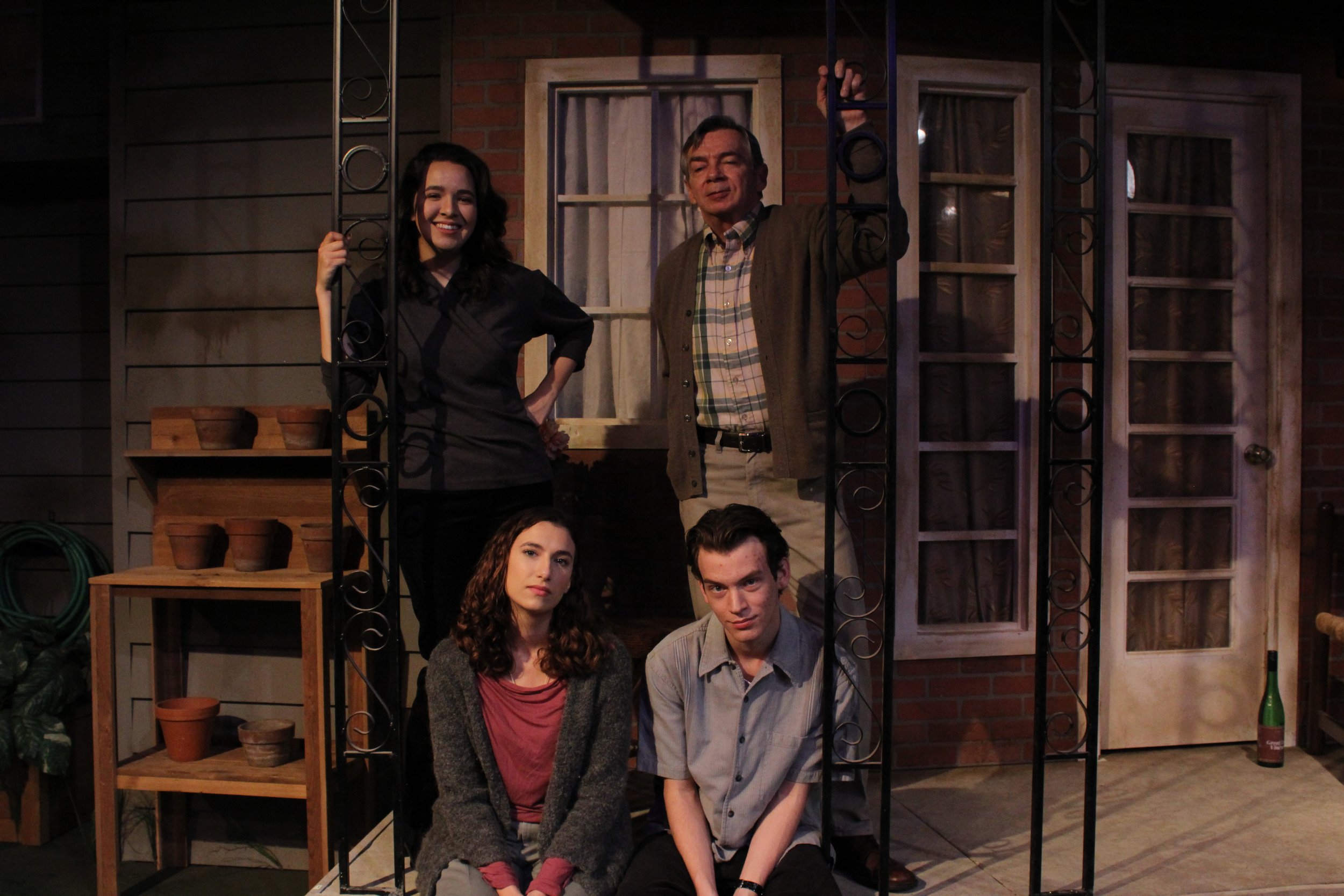 Four people posing on a porch. Two are standing, and two are sitting. Behind them are potted plants and a door with glass panels.
