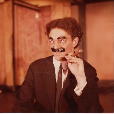 Person in a suit with glasses, mustache, cigar, and curly hair.