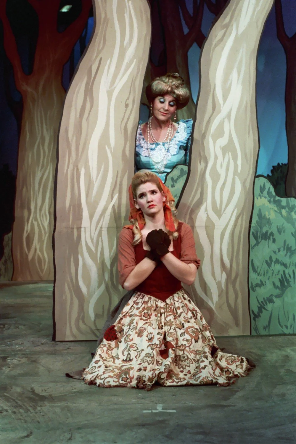 Two actors performing in a theatrical play set in a forest. One actor, with braided hair, kneels and appears pensive or distressed, wearing vintage-style clothing. The other actor, dressed in a blue dress with pearls and styled hair, looks through a 
