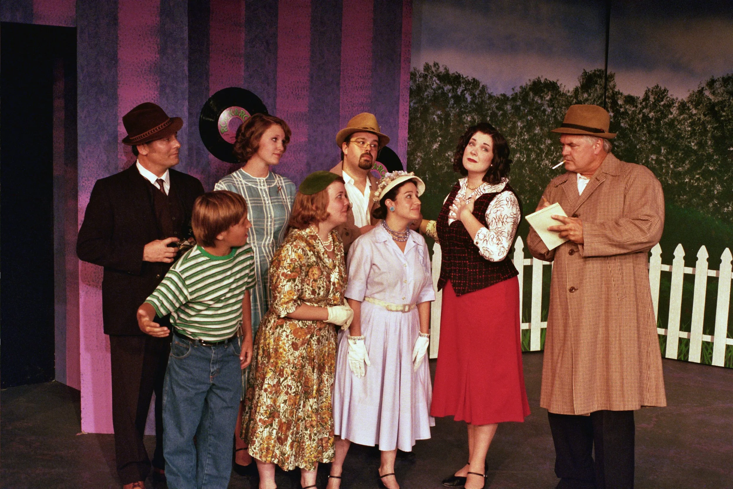 Group of people dressed in vintage 1950s clothing, on a stage set resembling a suburban scene with a painted fence and garden backdrop, similar to a community theater production.