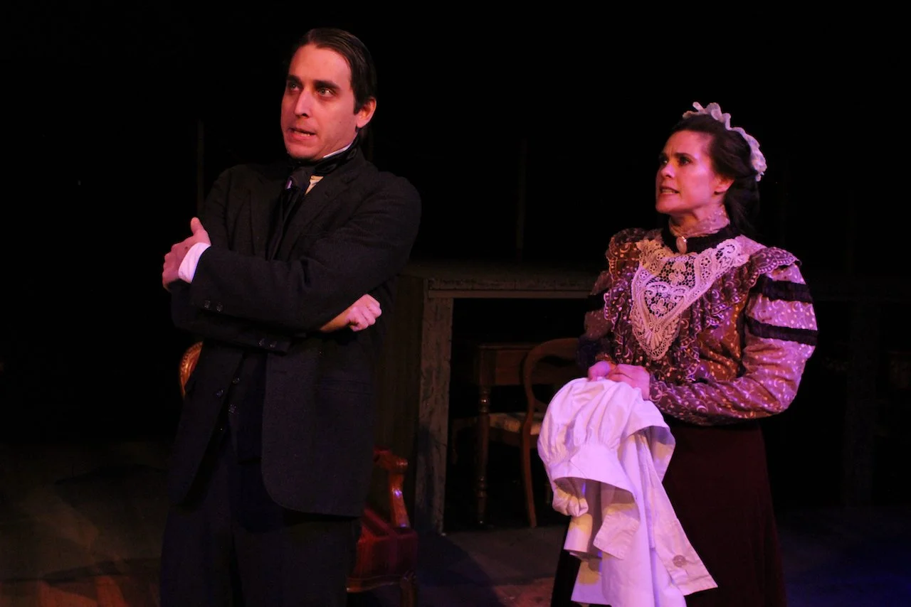 Two actors in period costumes performing on stage, one man standing with arms crossed and one woman holding a cloth, under stage lighting.