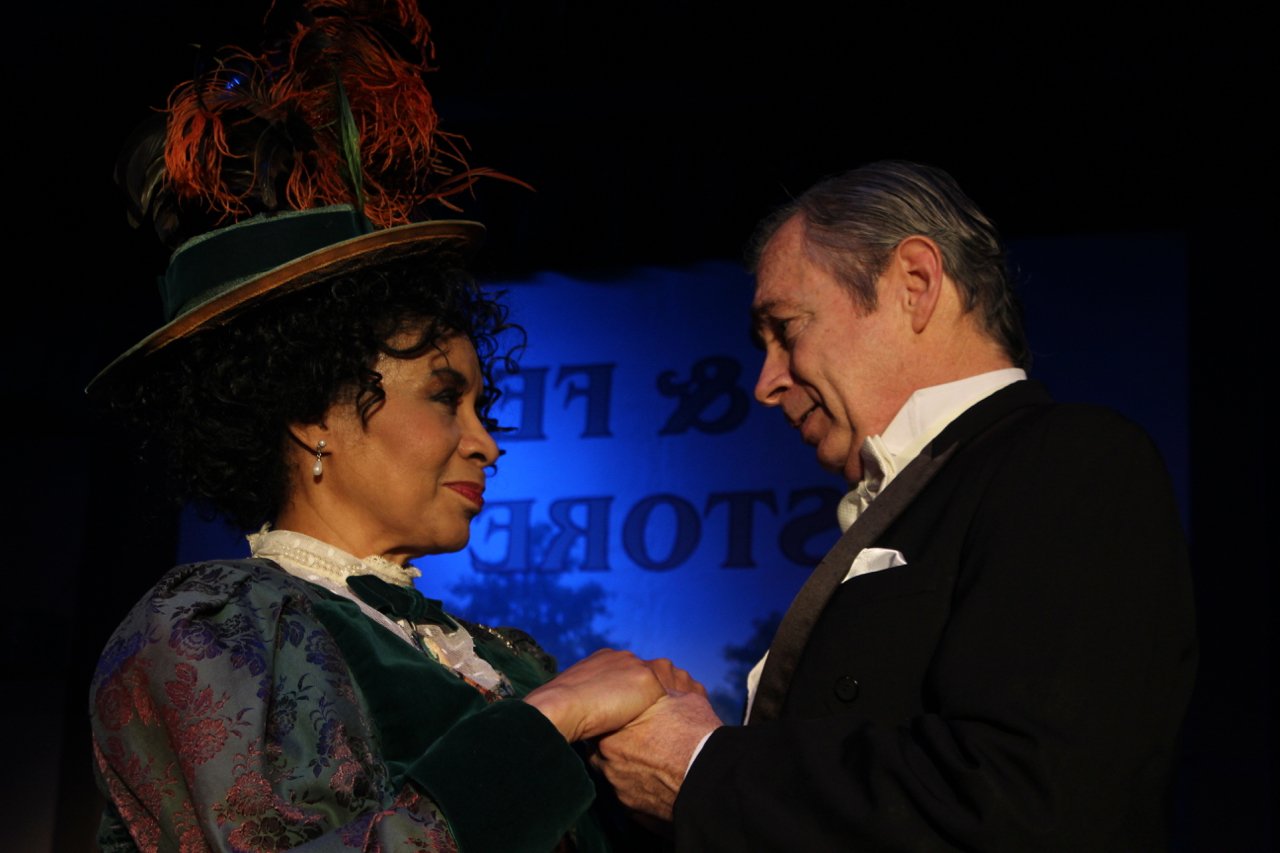 Two actors in period costumes perform on stage, wearing ornate outfits with a feathered hat and formal attire, holding hands intimately in a theatrical scene.