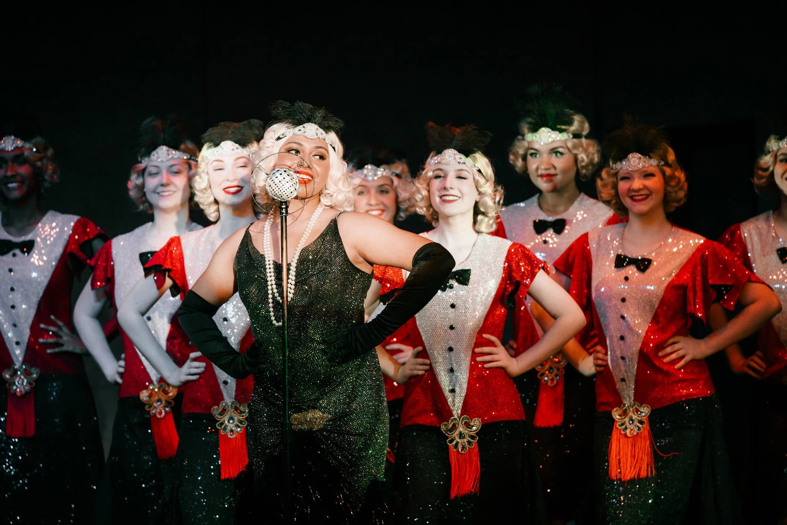 Performers in vintage costumes with blonde wigs and sequined dresses sing into a retro microphone on stage.