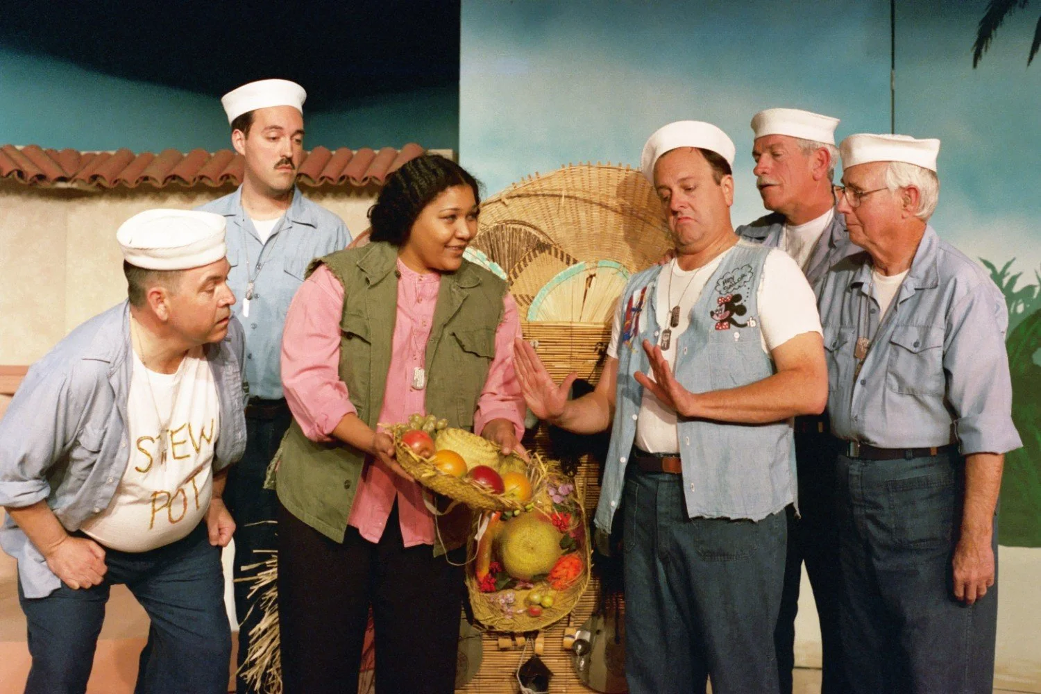 Group of actors dressed as sailors and a woman, holding a basket of fruit, performing a scene on stage.