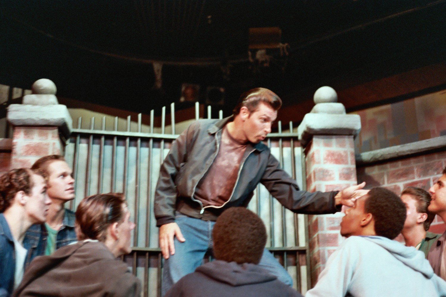 Group of men in front of a brick gate, one gesturing dramatically