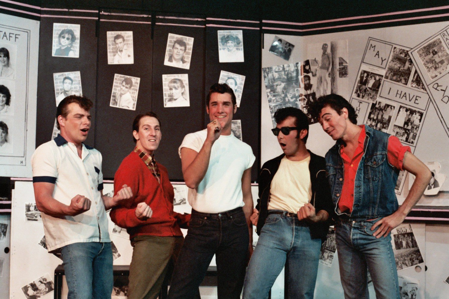 Five men performing in a retro-themed stage setting with black and white photo portraits and media on the wall.