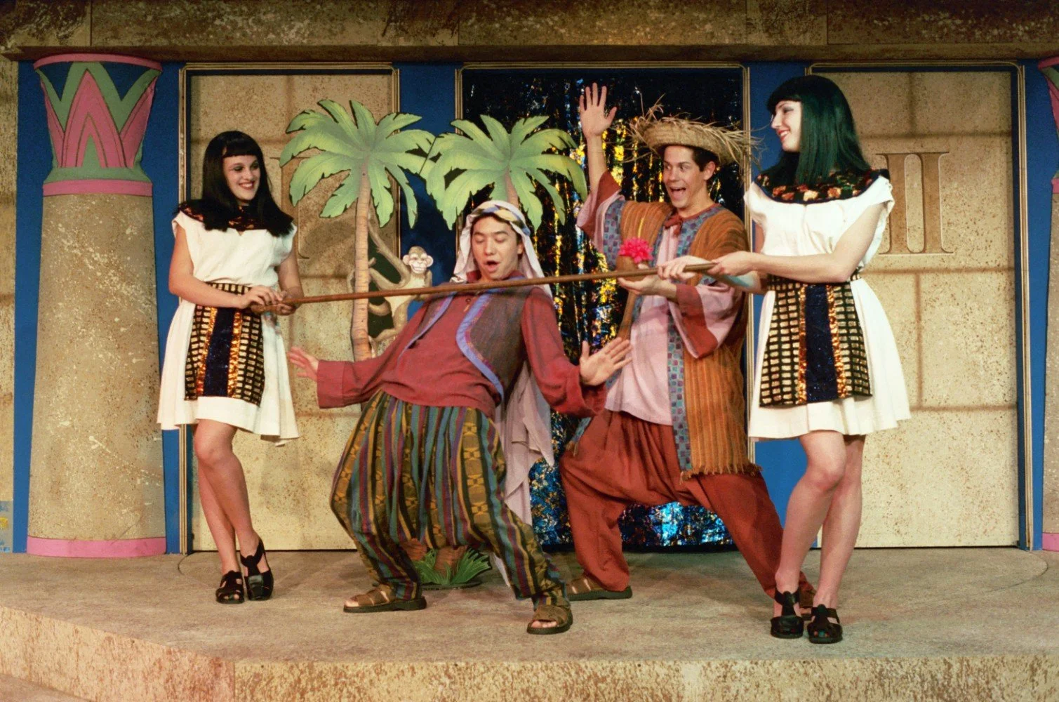Stage performance featuring actors in ancient Egyptian-style costumes, with a playful scene involving two women holding a stick while a man leans backward under it. A backdrop includes palm trees and a decorative Egyptian motif.