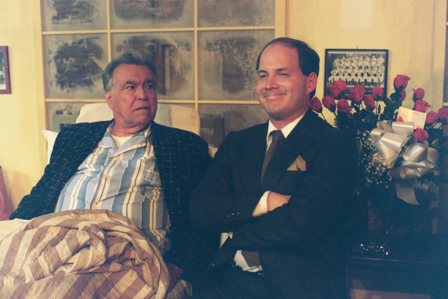 An elderly man wearing striped pajamas and a robe sits in a chair, next to a younger man in a suit. The younger man is smiling while the elderly man looks serious. In the background, there is a bouquet of red roses with a large silver ribbon.