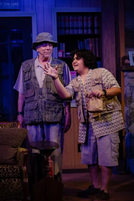 Two actors perform on stage in a room setting, one older wearing a bucket hat and vest, the younger with a patterned shirt and cargo pants. They appear engaged in conversation, possibly from a play or theatrical scene.