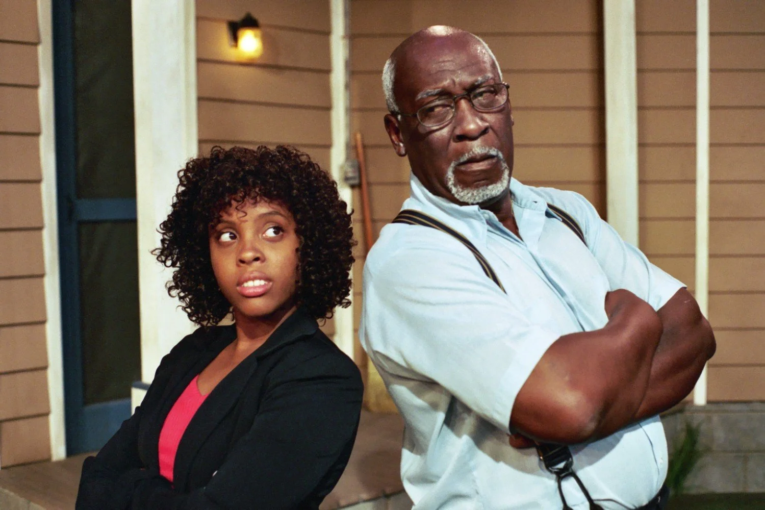 Two people standing back to back with arms crossed, one wearing a black blazer and curly hair, and the other wearing glasses and a light blue shirt, against the backdrop of a house with brown siding and a lit porch light.