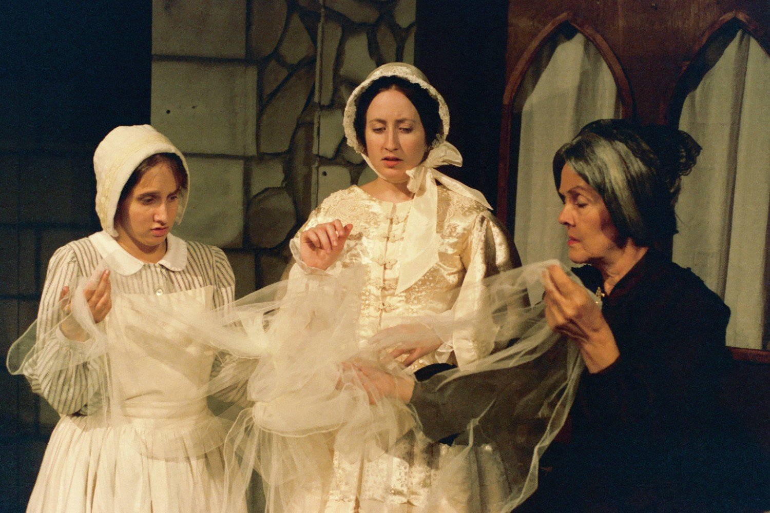 Three women in period costumes examining a delicate piece of fabric, likely in a theatrical setting with a stone wall and curtains in the background.