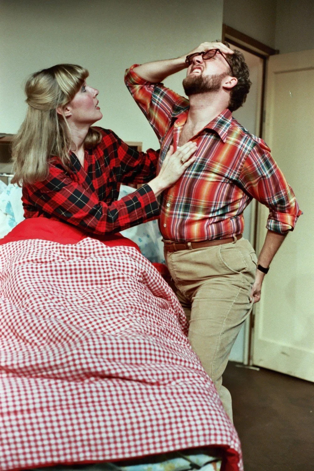 Two people in a dramatic scene; one person sitting on a bed, covered with a red checkered blanket, and the other standing, both wearing plaid shirts, expressing strong emotions.
