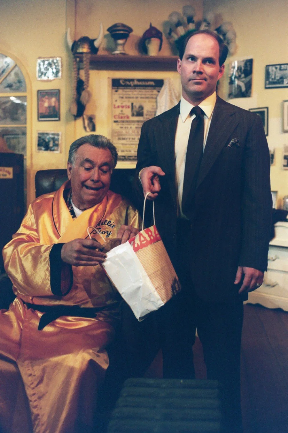 Two men in a room; one in a yellow robe and the other in a suit, holding a white and red shopping bag. The room has a vintage decor with various hats and framed photos on the wall.