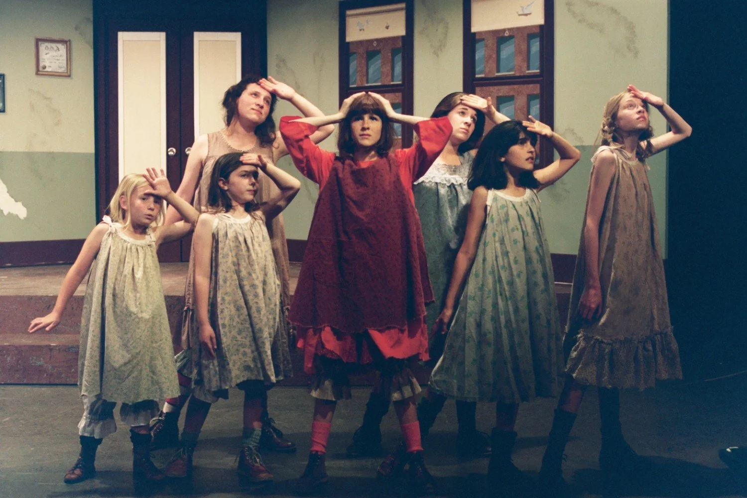 Group of girls and a woman on stage during a theatrical performance. They are wearing nightgowns and looking upwards with hands shading their eyes. The background resembles an interior setting with doors and windows. The scene suggests a moment of di