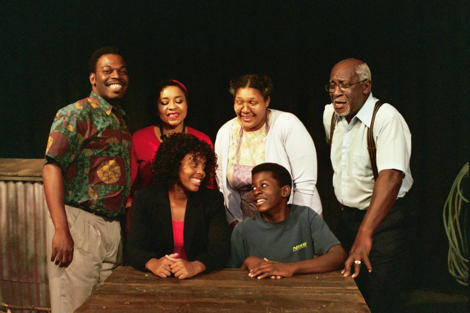 Group of six people smiling around a table in a dimly lit room, with one person seated and the others standing.