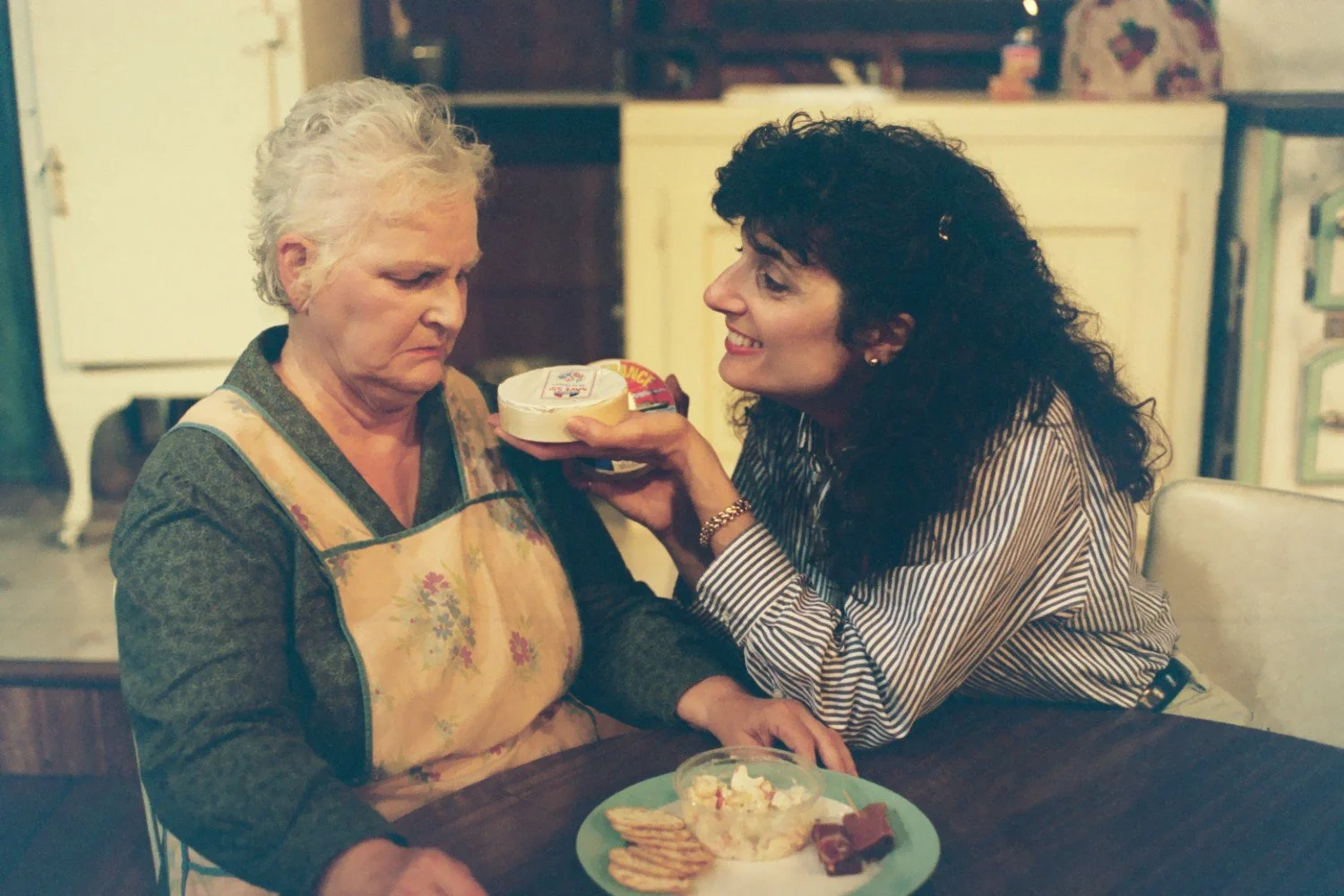 Two women at a table; one holds a container of food, while the other, wearing an apron, looks unimpressed. On the table, there's a plate with crackers and a small bowl of salad.