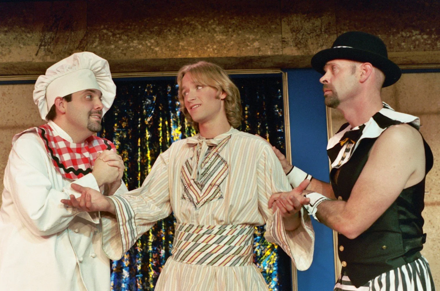 Three people in theatrical costumes, including a chef, standing on stage with expressions of engagement.