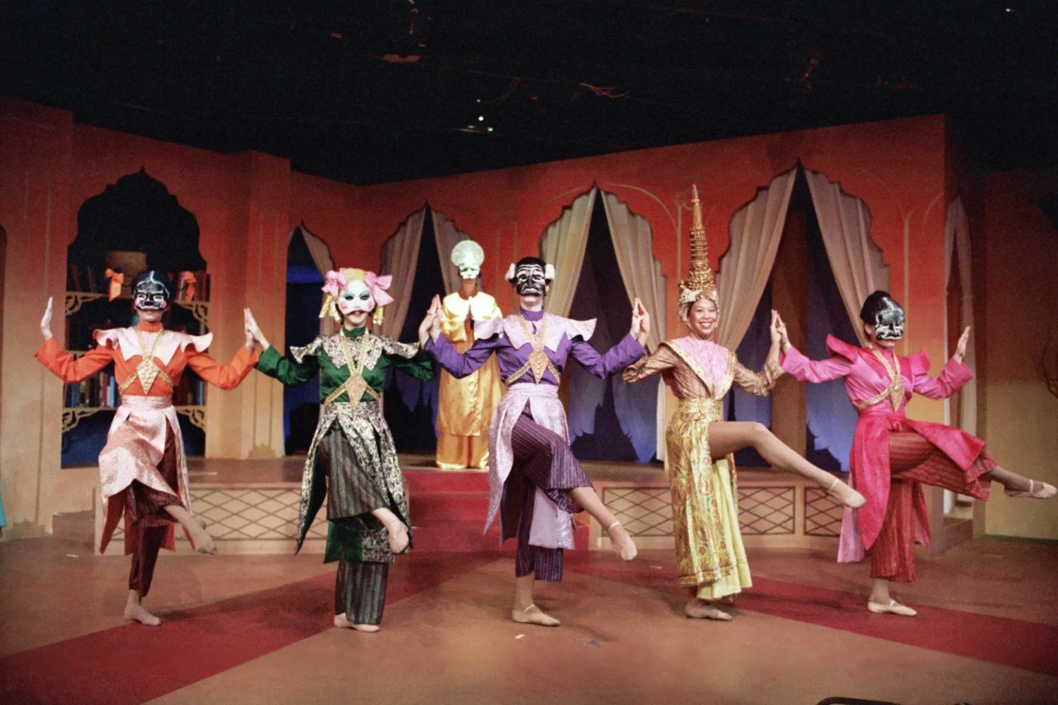 Group of performers in traditional Asian costumes and masks on stage, posed in a dance formation.