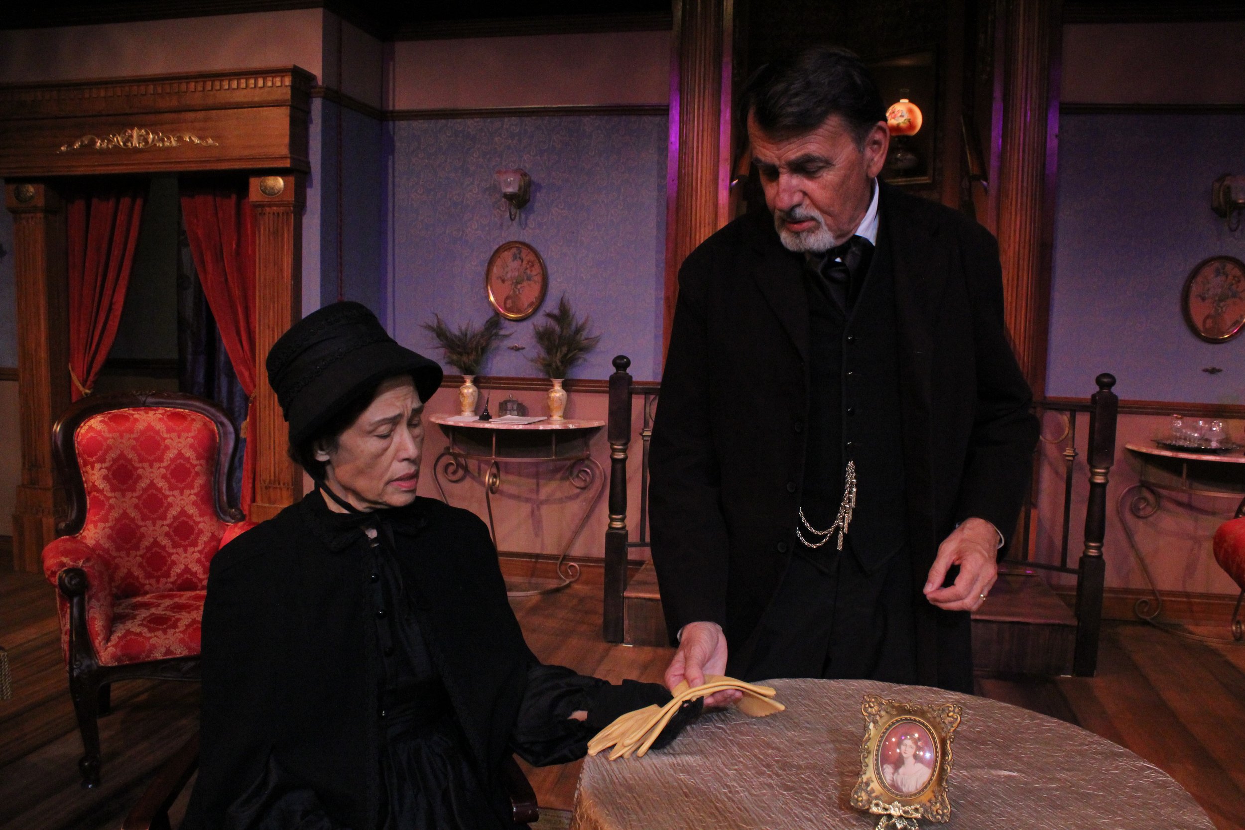 Two actors in period costumes in a Victorian-style room with a framed photo on the table, depicting a dramatic scene.