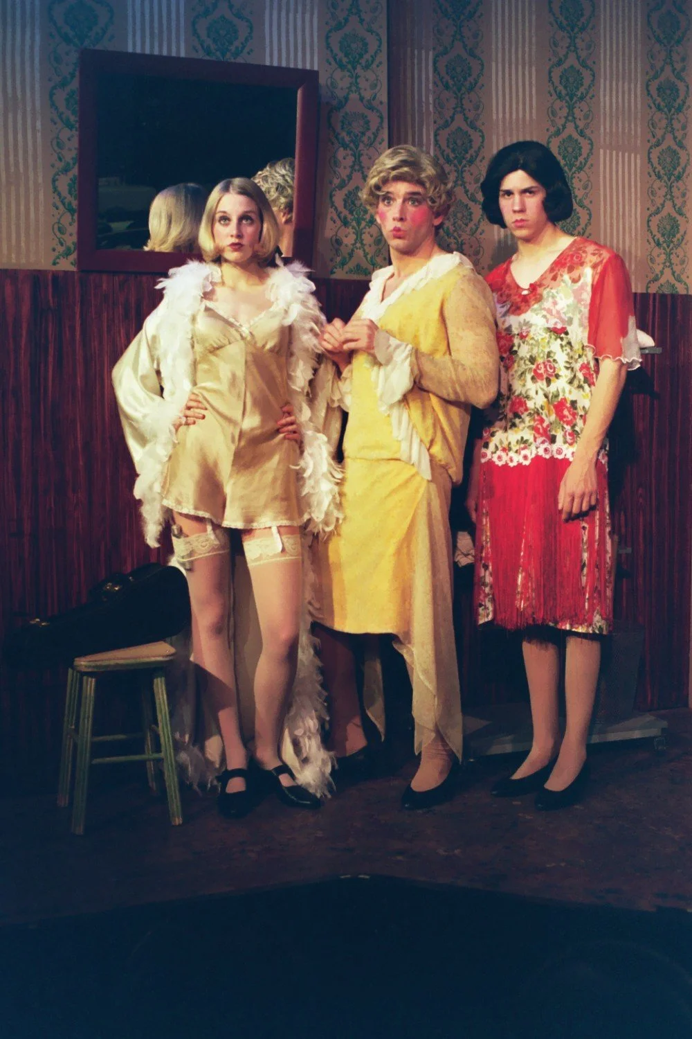 Three people wearing vintage-style dresses in a room with retro wallpaper, including two men dressed in women's clothing. A mirror is on the wall and a stool with a violin case is in the foreground.