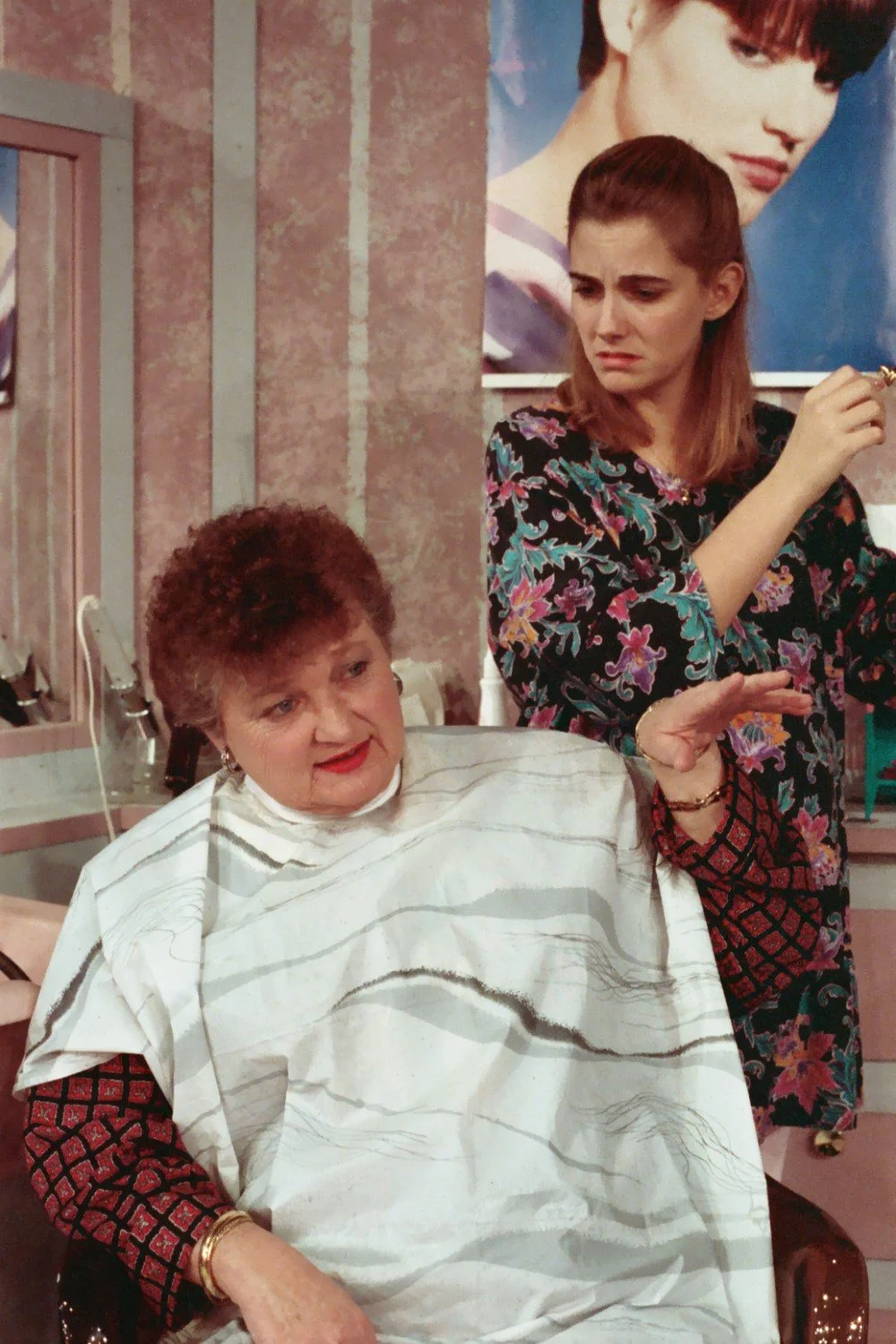 Two women in a hair salon, one seated in a salon cape, the other standing with scissors, looking perplexed. There's a poster of a hairstyle on the wall behind them.