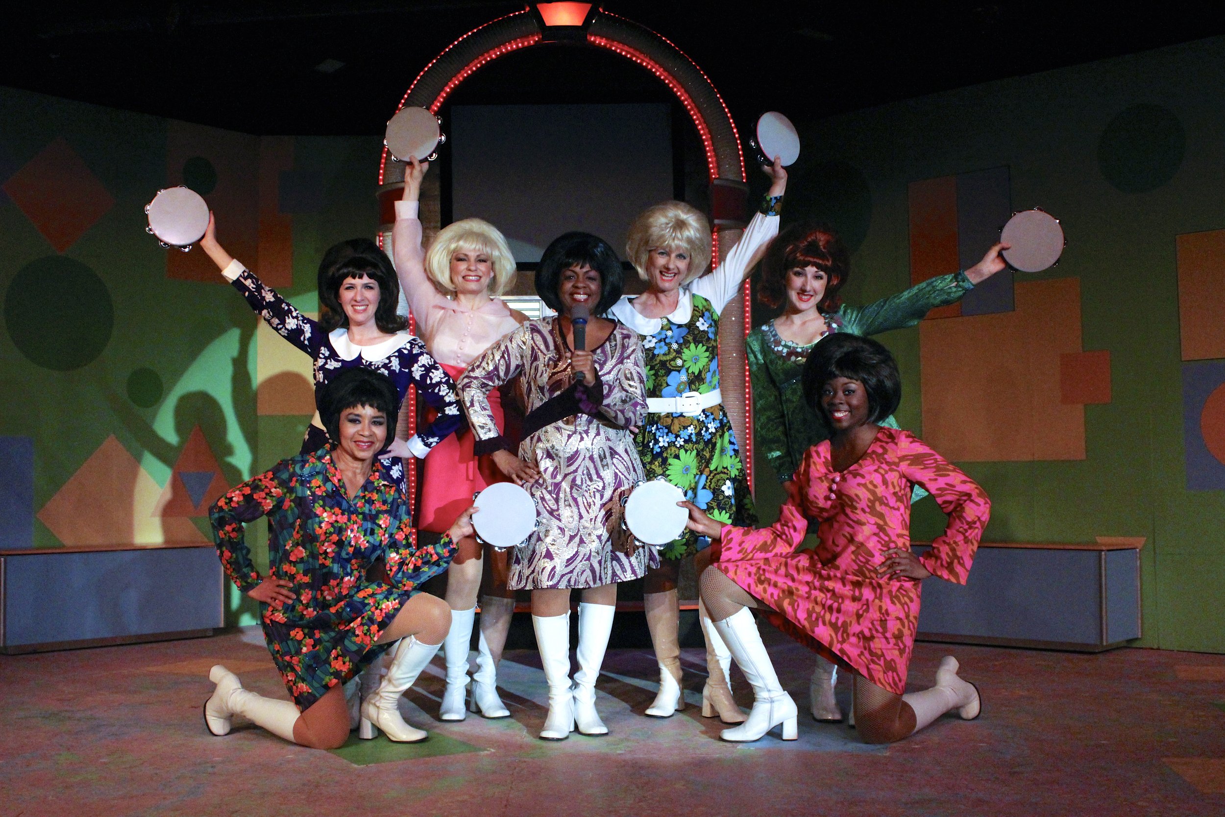 Seven women in colorful 1960s-style dresses and white boots, posing with tambourines on a stage with a retro geometric background.