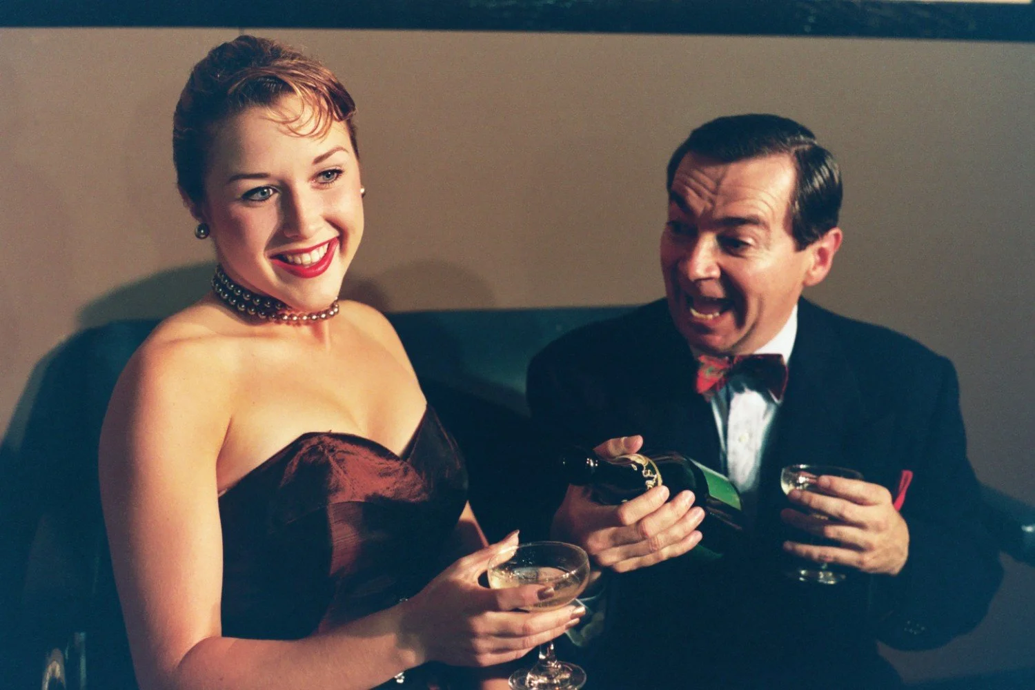 A woman in a red dress smiling while a man in a suit opens a bottle of champagne, with both holding glasses.