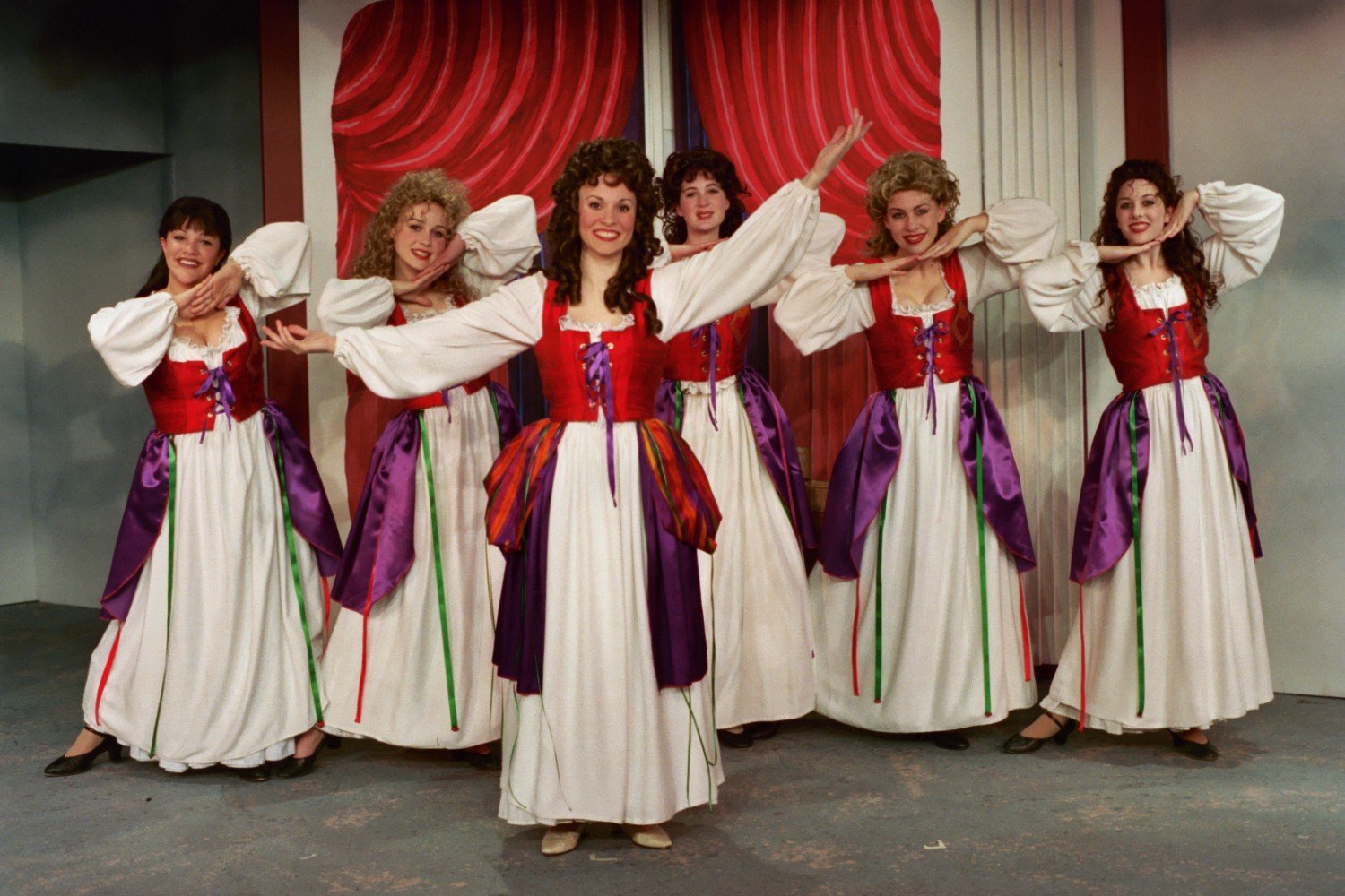 Six women dressed in period costumes with white dresses, red bodices, and purple sashes, posing in front of a red curtain.