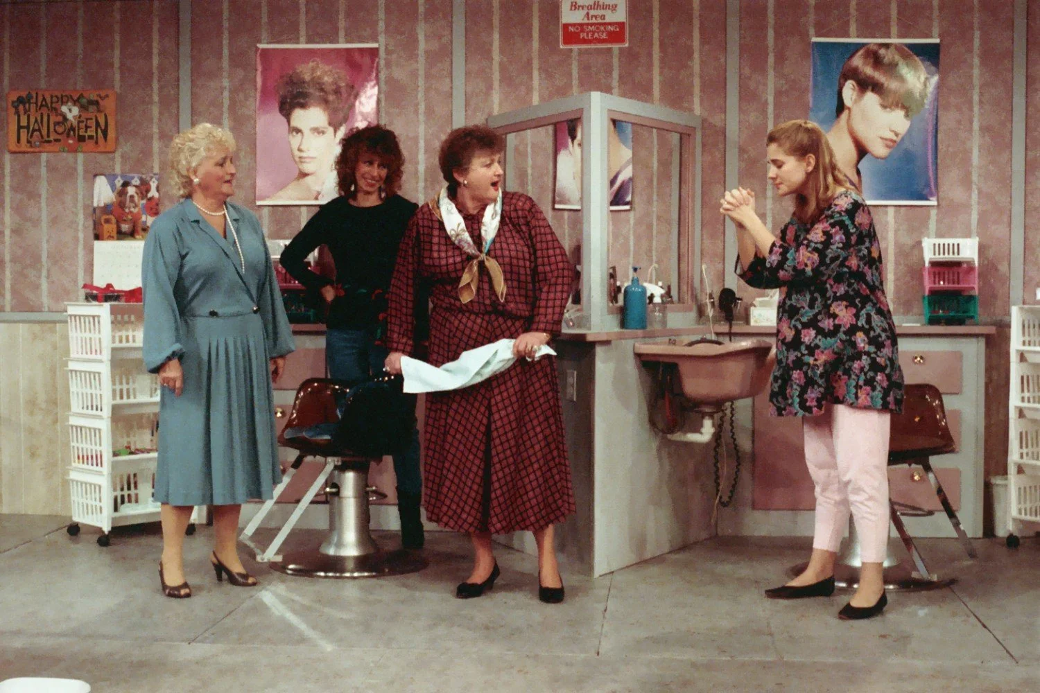 Scene inside a hair salon with four women talking, two hairdresser chairs, hair products, and hair posters on the walls. A Halloween decoration is visible.