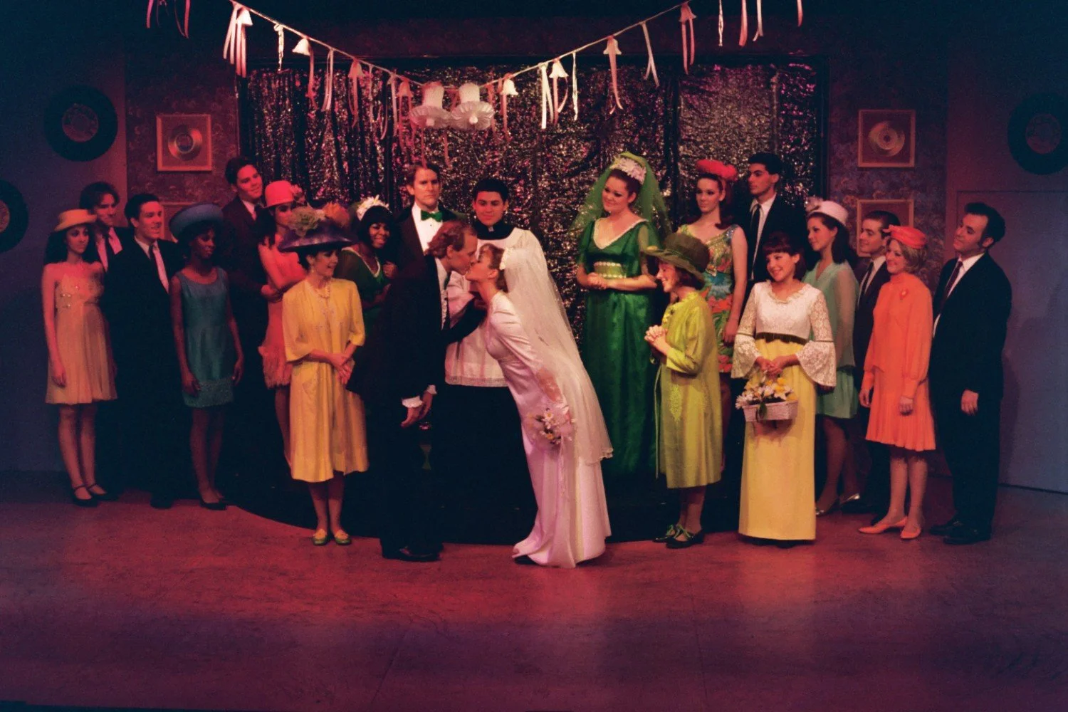 A stage performance with actors in a wedding scene, featuring a bride and groom kissing. The ensemble is dressed in colorful retro attire, with women in bright dresses and hats, and men in suits, set against a backdrop with ribbons and a sparkly curt