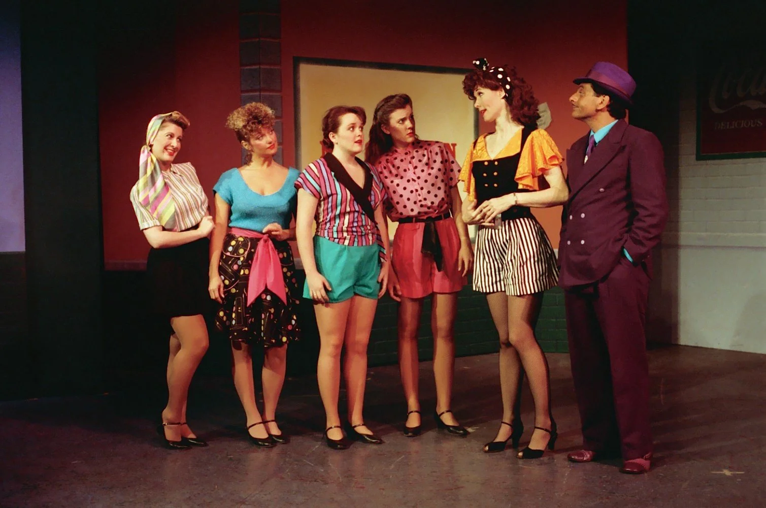 Group of performers in colorful 1980s attire on stage with a Coca-Cola sign in the background.