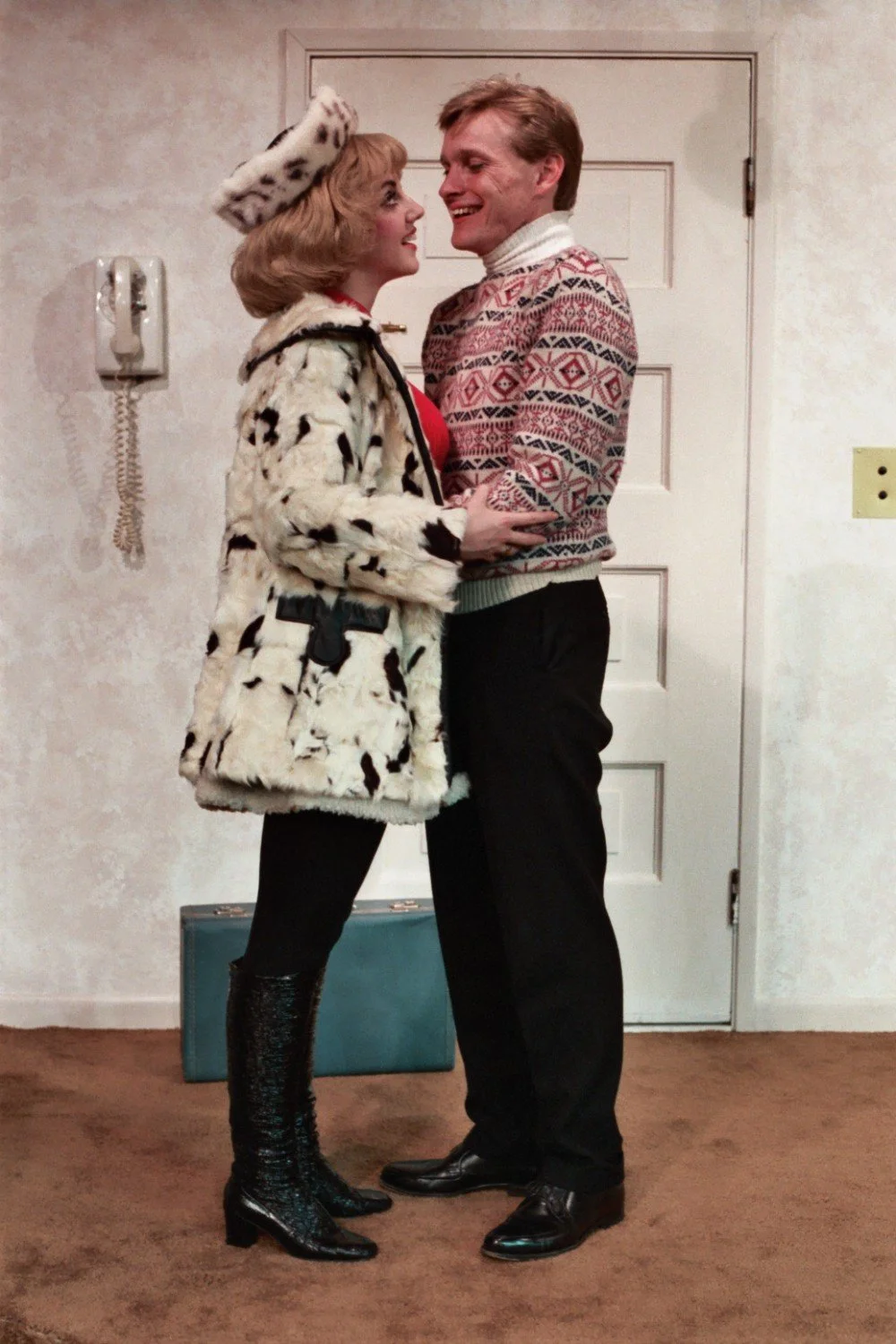 A man and woman dressed in winter clothing smiling at each other inside a room. The woman is wearing a spotted coat and matching hat, while the man is wearing a patterned sweater. They stand near a door with a wall-mounted phone visible in the backgr