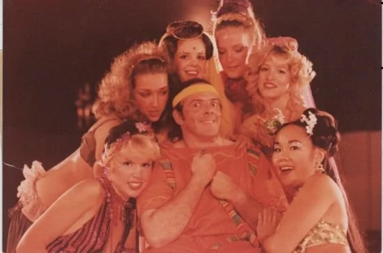 A man in a colorful costume and headband surrounded by six women in vibrant, embellished attire and elaborate hairstyles, posing together in a playful manner.
